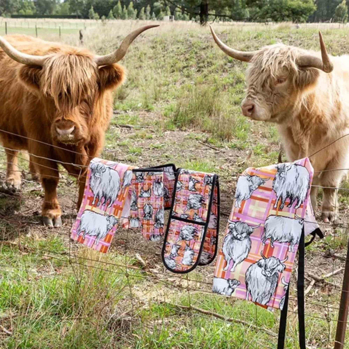 Highland Cows double oven glove featuring illustrated Highland cows on a pink, orange and cream tartan background with quilted heat-resistant cotton construction. Also in image are matching oven glove and pot holder set, tea towel and apron.