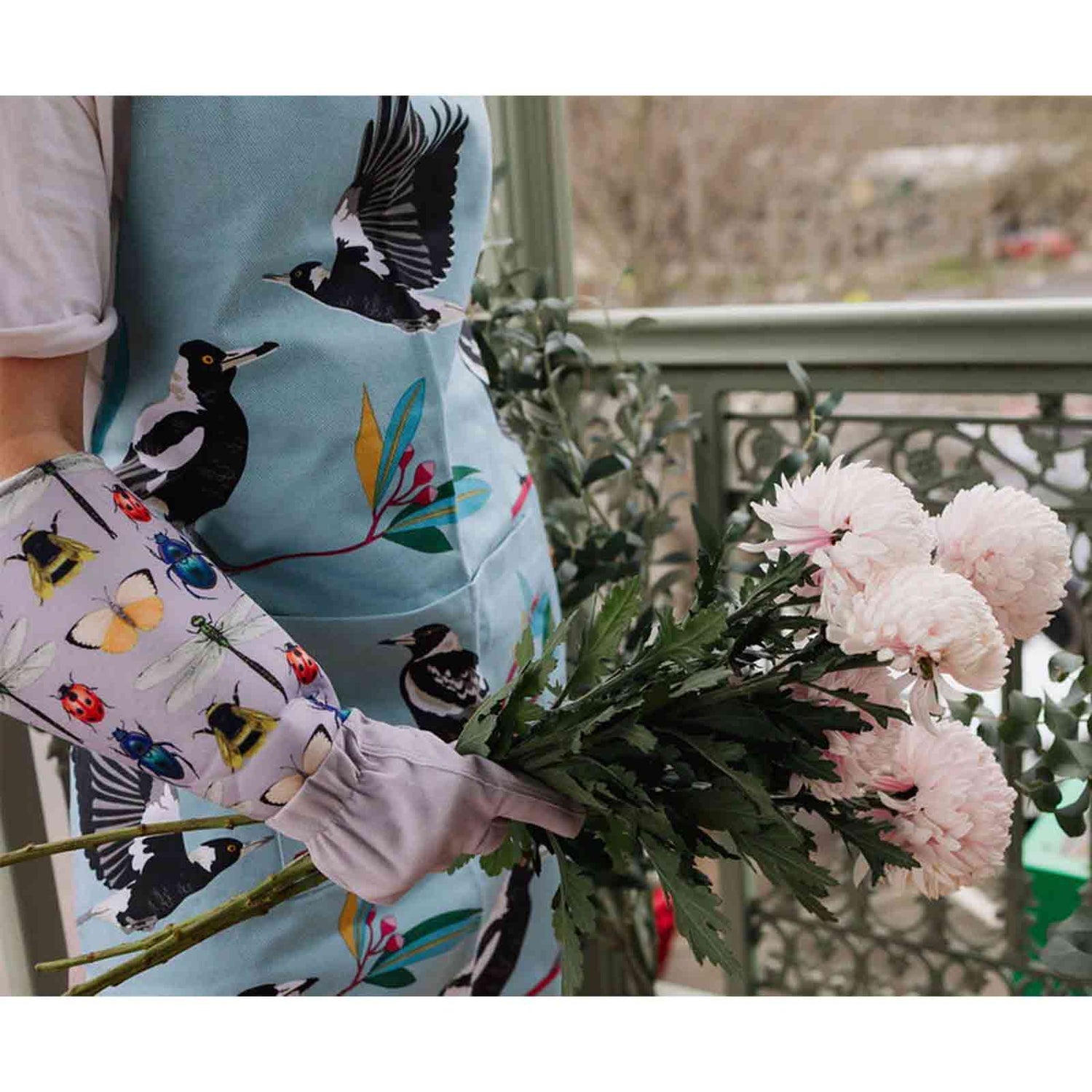 Person holding a bouquet of flowers with a colorful apron featuring bird designs and gardening gloves with an insect design