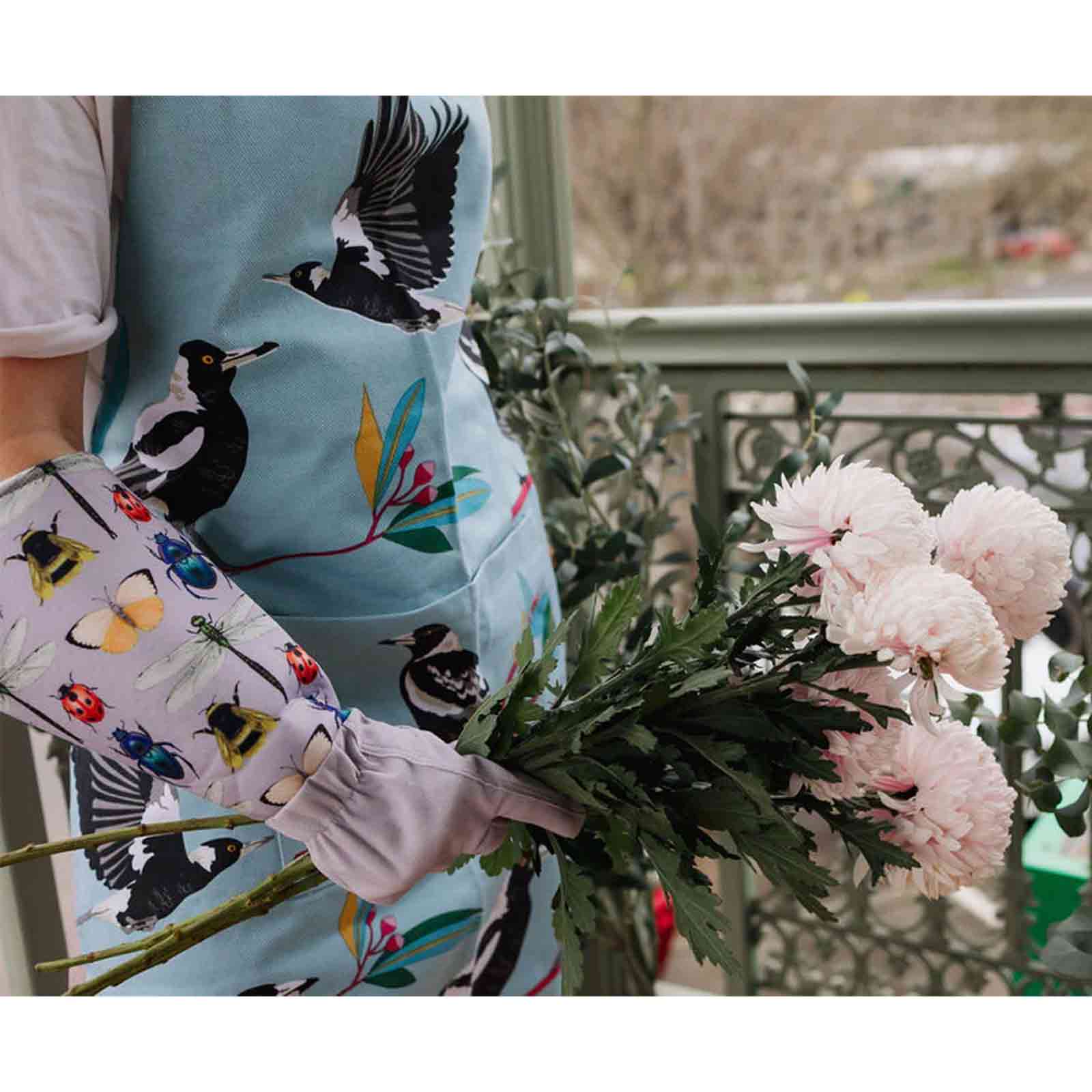 Person holding a bouquet of flowers with a colorful apron featuring bird designs and gardening gloves with an insect design