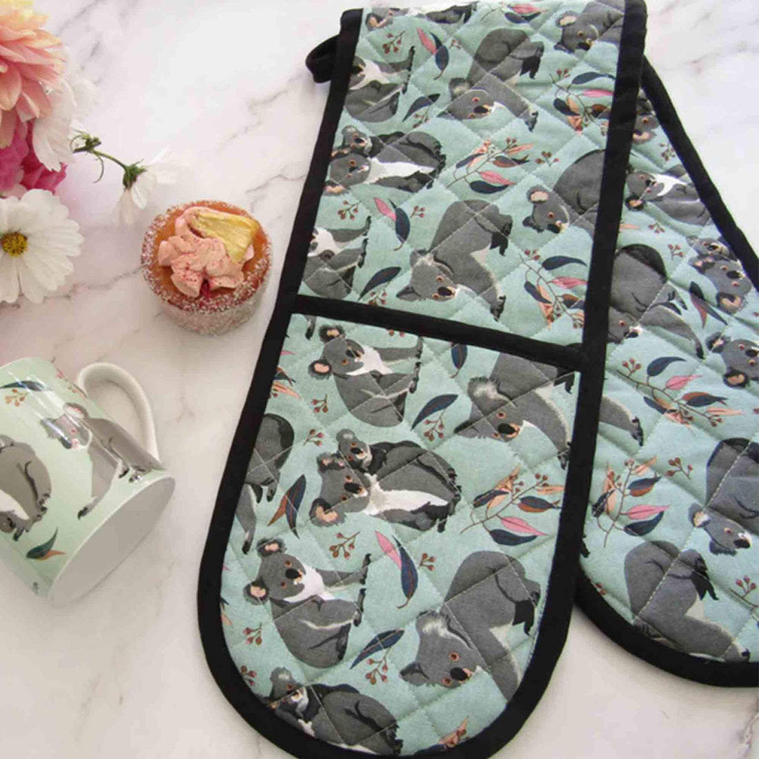 Oven mitts with koala design on a marble surface with a cupcake and mug.