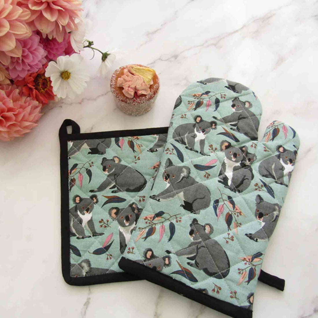 Set of pot holder and oven mitt with koala design on a marble surface with flowers and cupcake.