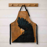 Brown apron with a Black Cockatoo design hanging on a wooden rack against a white wall.