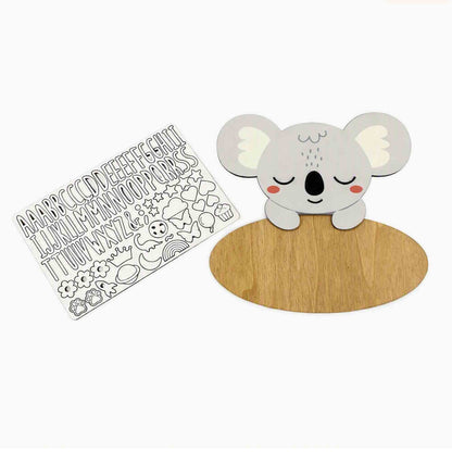 A wooden koala-themed name sign and matching alphabet sticker sheet on a plain white background. The koala is light gray with white ears, a black nose and mouth, pink cheeks, and sits on a round natural wood base.
